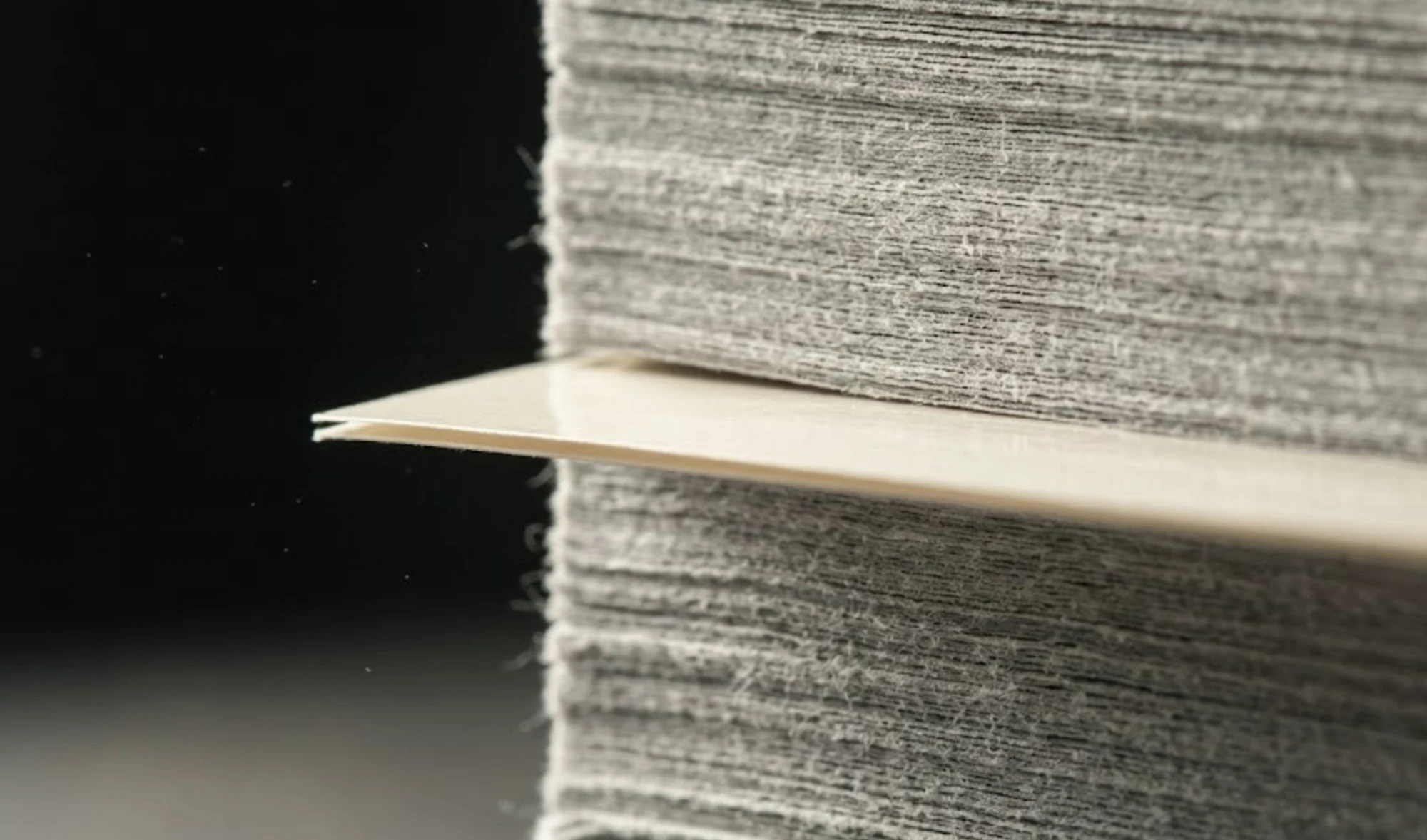 Detailní snímek stohu novinového papíru, mezi jehož vrstvami je vsunutý jediný hladký lesklý list, který tam nepatří.